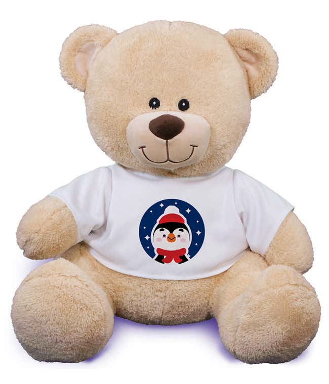 Tan stuffed teddy bear wearing a white t-shit that has a characterized penguin in a scarf and hat on it.